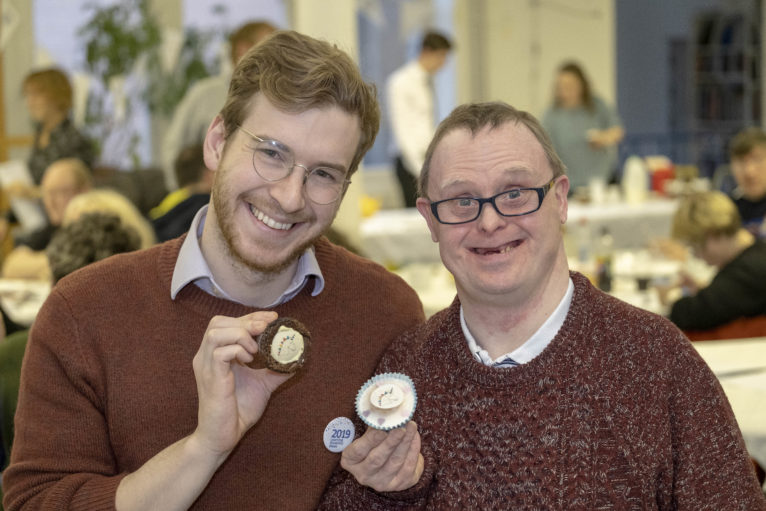 celebrity baker James Morton sits beside Stephen Dickson who designed the Learning Disability Week mascot, 'Uno the Unicorn', both holding cup cakes with Uno the Unicorn cake toppers on them.