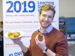 James Morton holds a plate of cake and takes a bit out of his banoffee traybake cake whilst standing in front of a learning disability week 2019 pop up banner