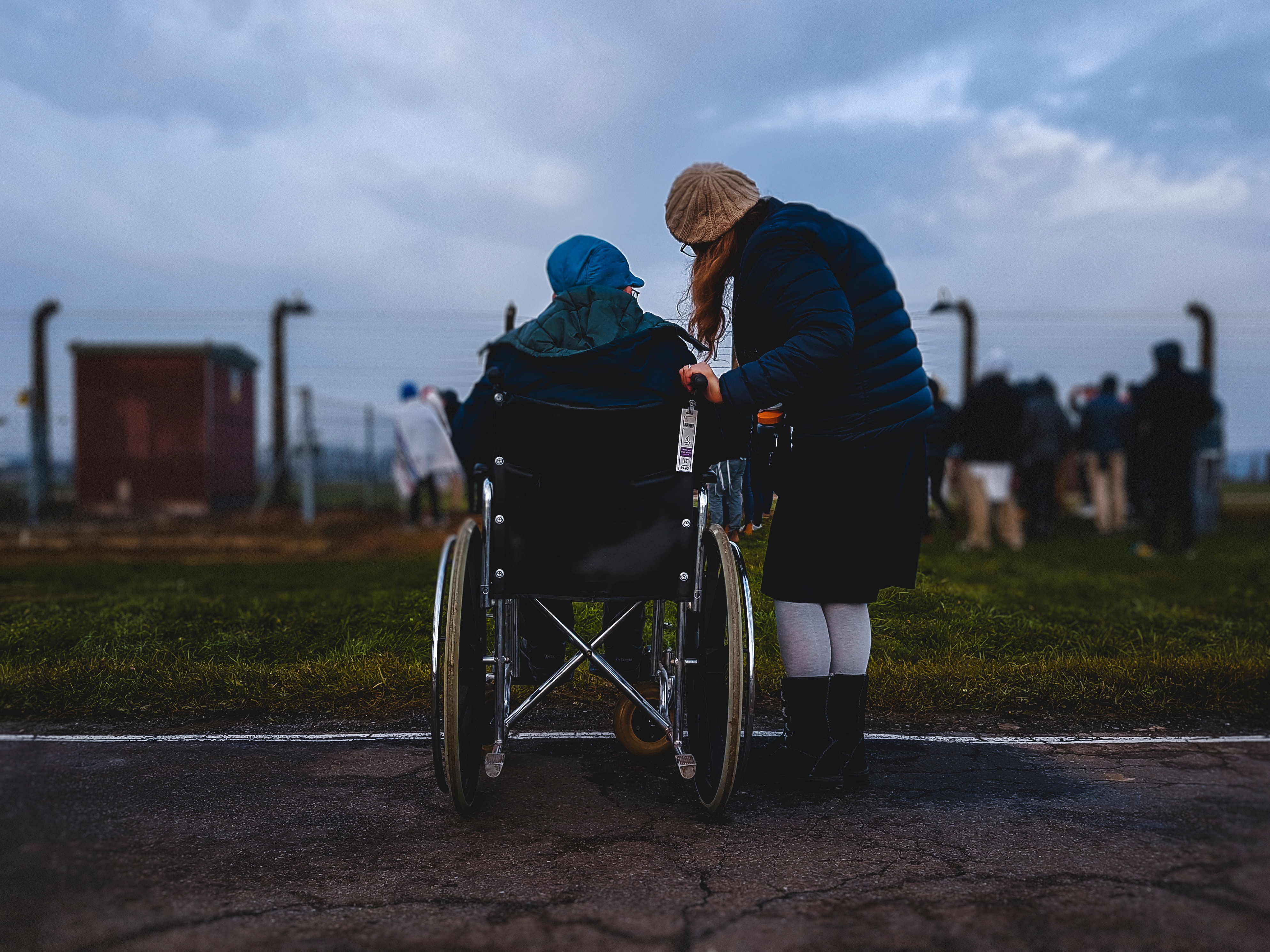 Person in a wheelchair sits looking out at field, beside them is a woman speaking to them.