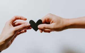 Two people hold a small black paper heart between their hands. Photo credit: Kelly Sikkema on Unsplash.